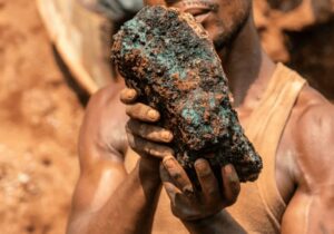 A man in a muddy tanktop holds a large chunk of greenish stone.