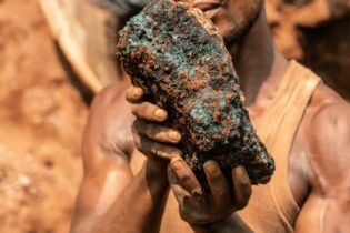 A man in a muddy tanktop holds a large chunk of greenish stone.
