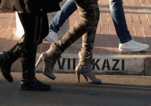 Low-level view of legs and feet of people walking along a street; painted street name visible on the kerb